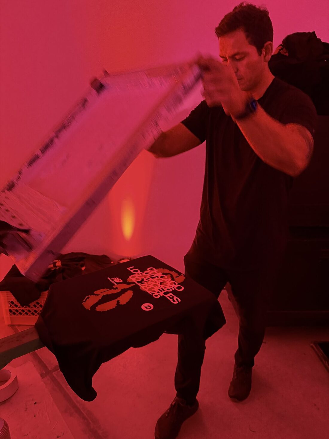 Printer lifting a screen to reveal a freshly printed black shirt under red party lighting