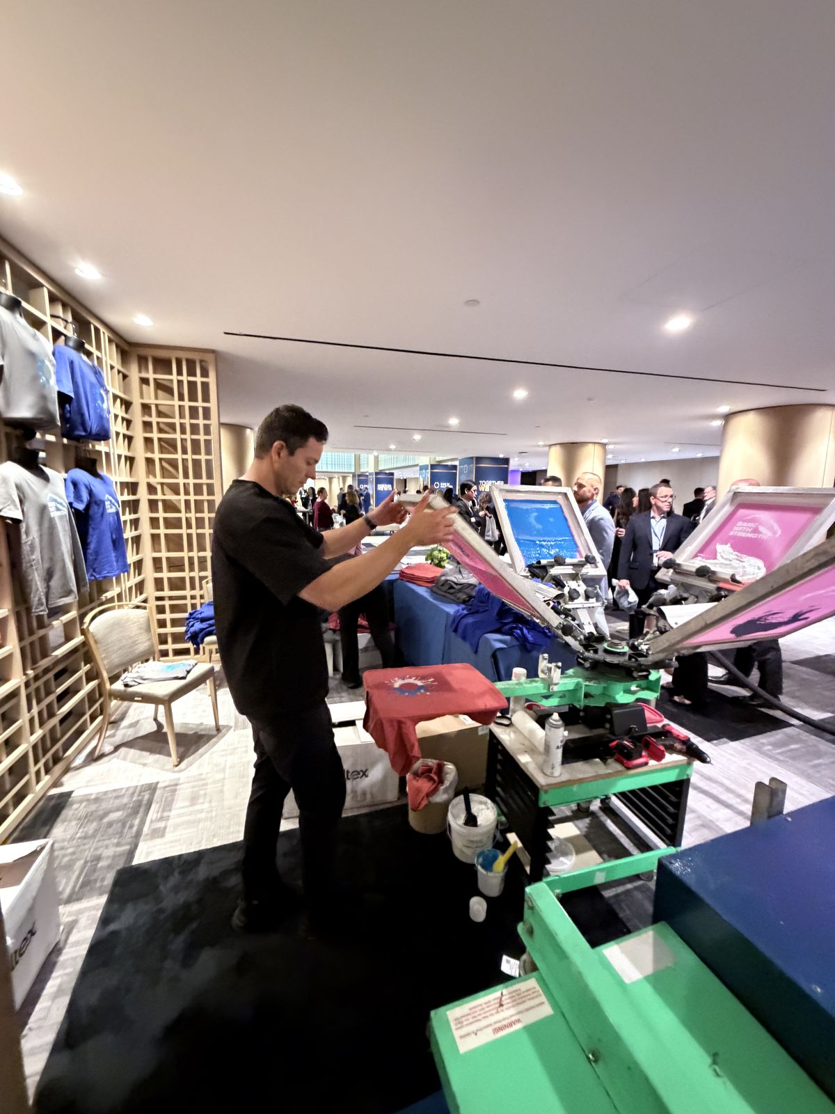 Merch Troop printer operating a multi-station press surrounded by folded apparel at a live event